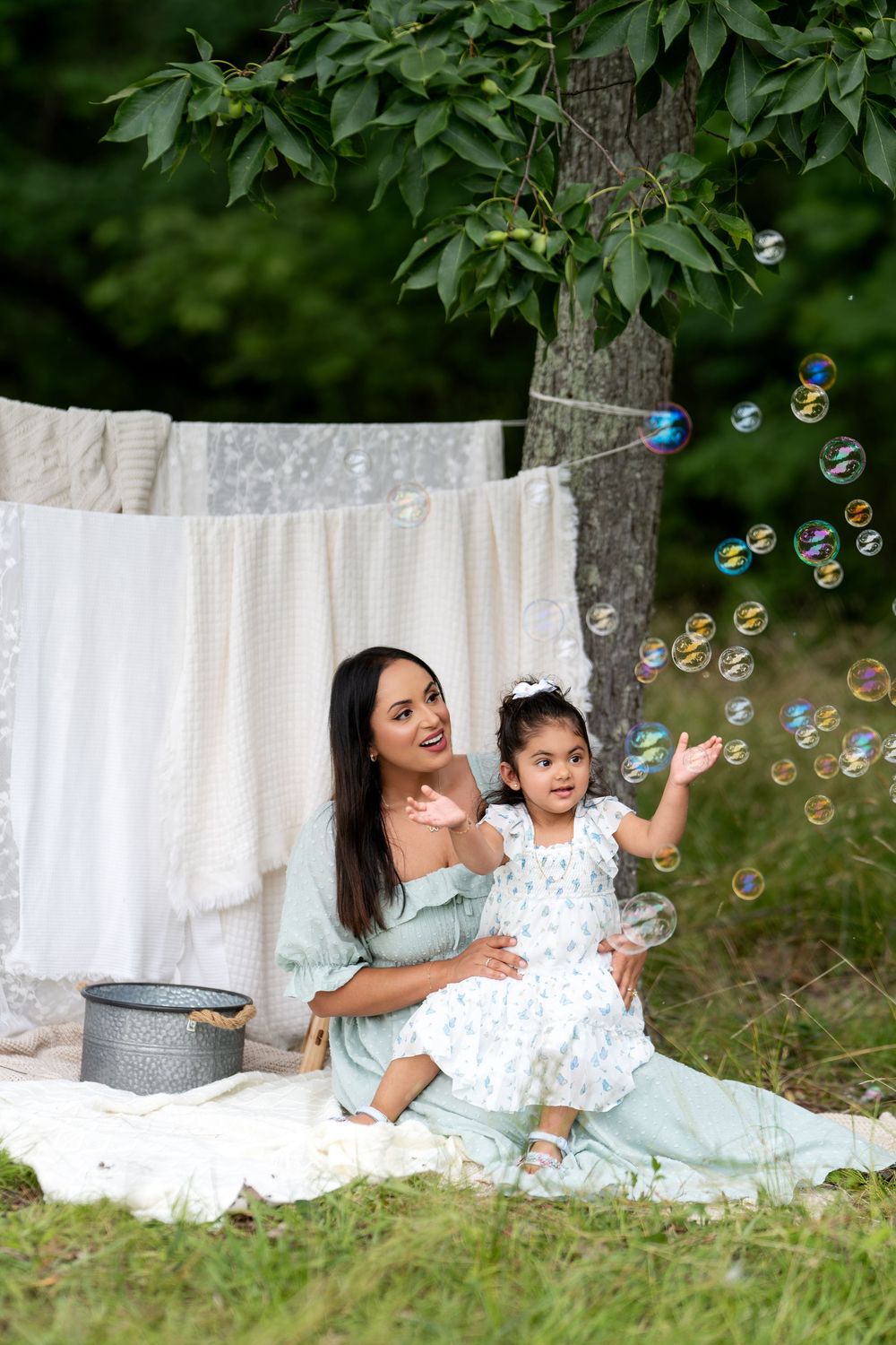 A mother and young daughter sit on a blanket outdoors, laughing and reaching for colorful soap bubbles near a clothesline.
