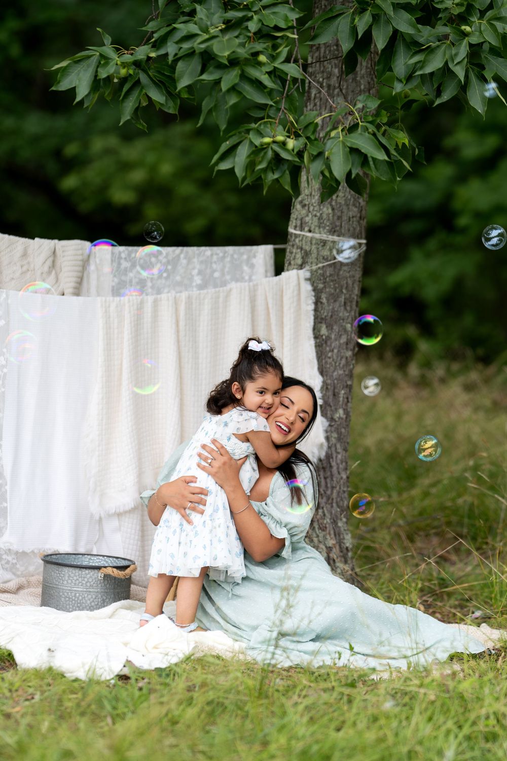 A mother and young daughter laugh and hug outdoors near a tree with white linens hanging, surrounded by floating soap bubbles.
