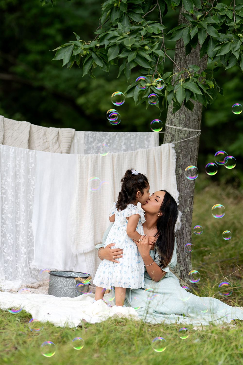 Mother and daughter share a tender kiss outdoors, surrounded by soap bubbles near hanging white linens in a lush garden.