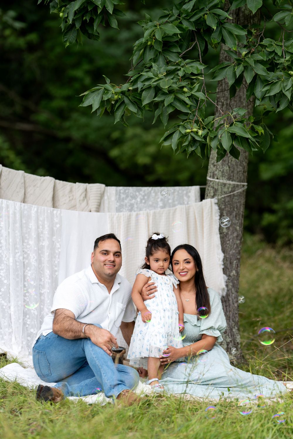 Family of three posing outdoors on a blanket near hanging white linens between trees in a lush green setting.