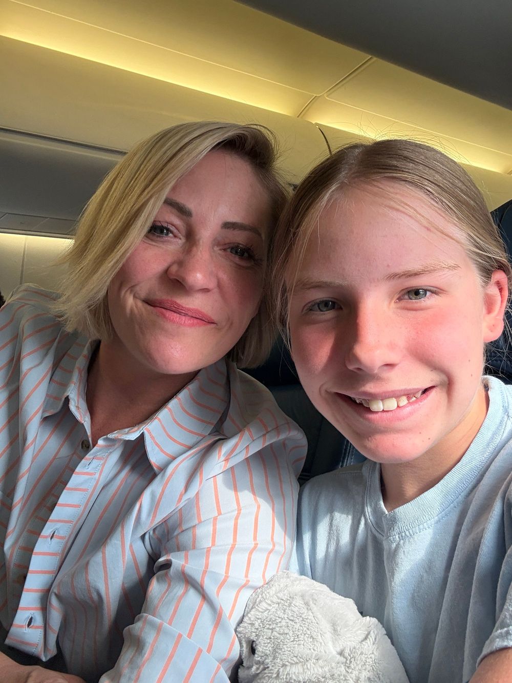 A woman and young girl smiling together for a selfie on an airplane, both wearing light blue tops.