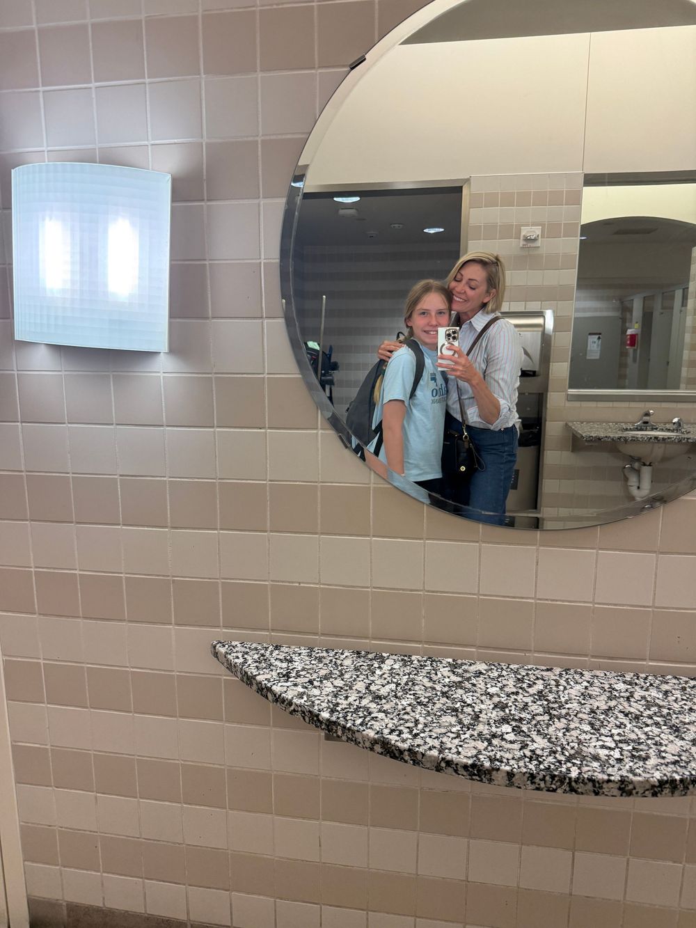 Two women taking a mirror selfie in a modern bathroom with round mirror, granite countertop, and beige tile walls.
