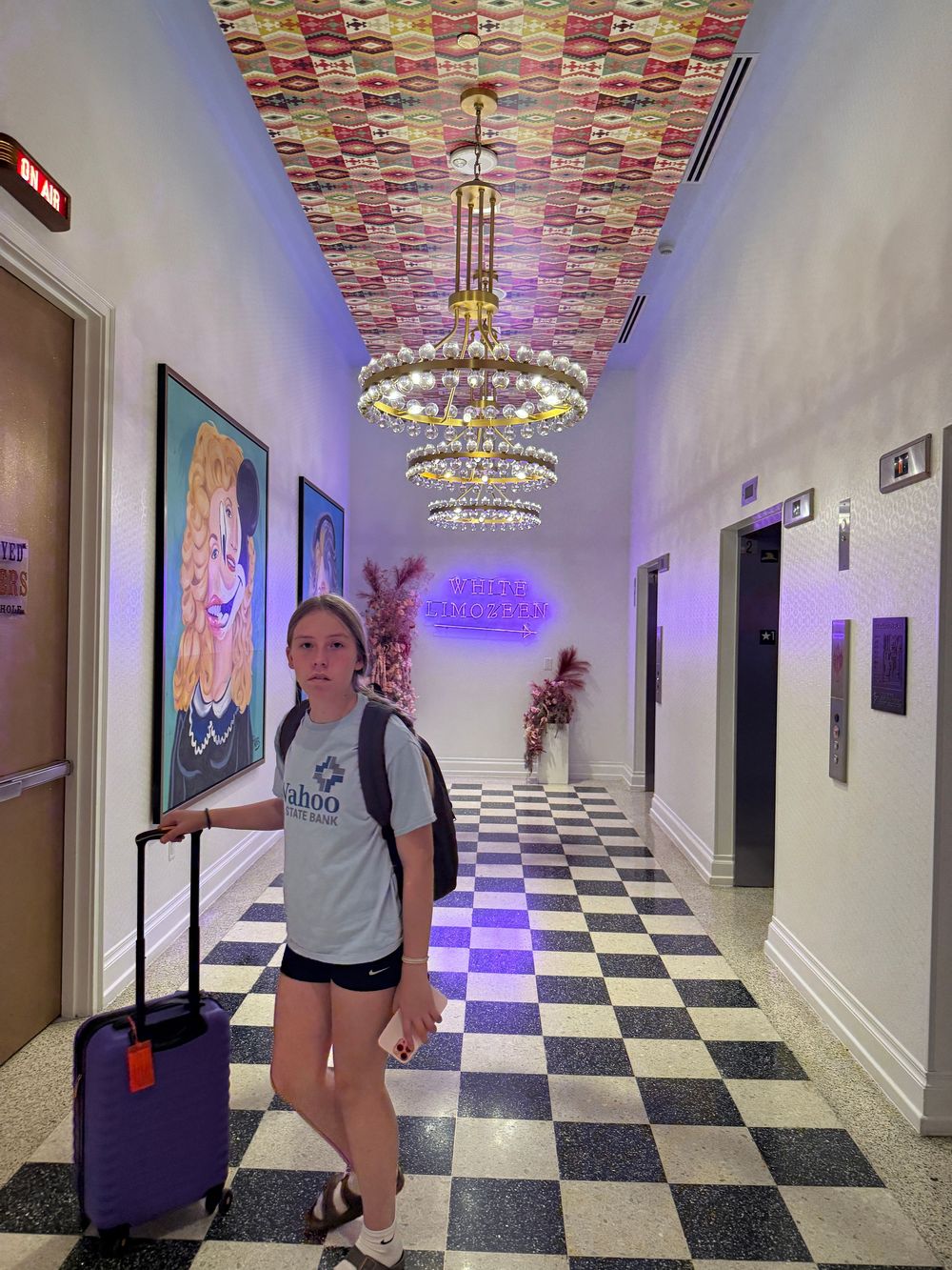 Girl with purple suitcase stands in decorative hotel hallway with chandelier, checkered floors, and colorful ceiling art.
