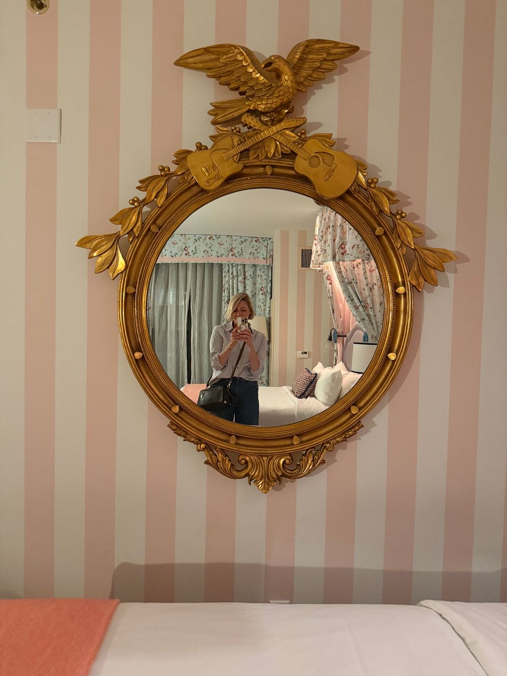 Ornate gold eagle mirror with round frame on pink striped wall, person taking selfie reflected in bedroom.