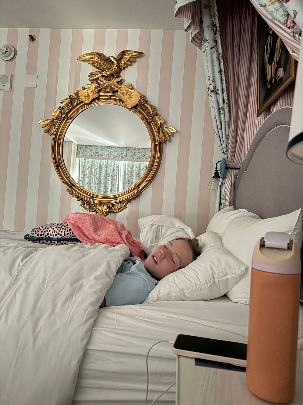 Child sleeping in bed beneath ornate gold eagle mirror on pink striped wall with white bedding.