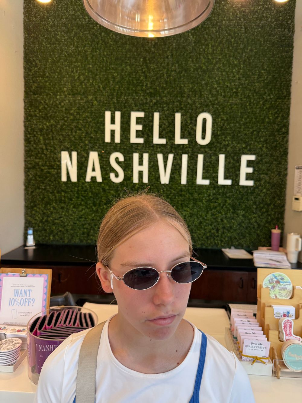 Young woman wearing oval sunglasses poses in front of a Hello Nashville green wall sign in a gift shop.
