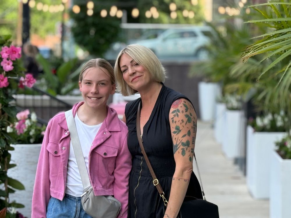 Two women smiling together outdoors at a patio venue, one in a pink jacket and one with a floral sleeve tattoo.