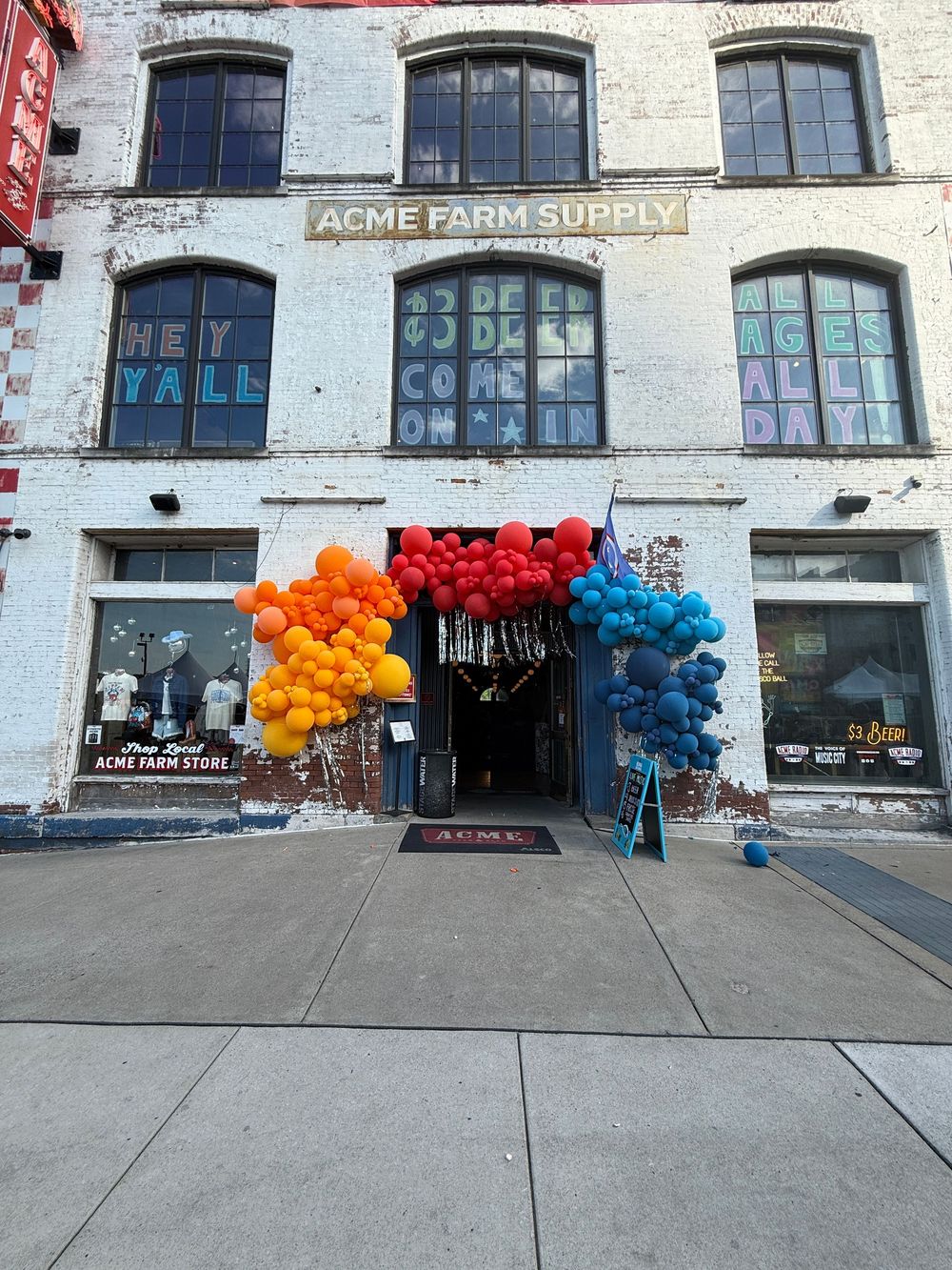 Colorful balloon arch decorates the entrance of Acme Farm Supply store on a brick building with festive window displays.