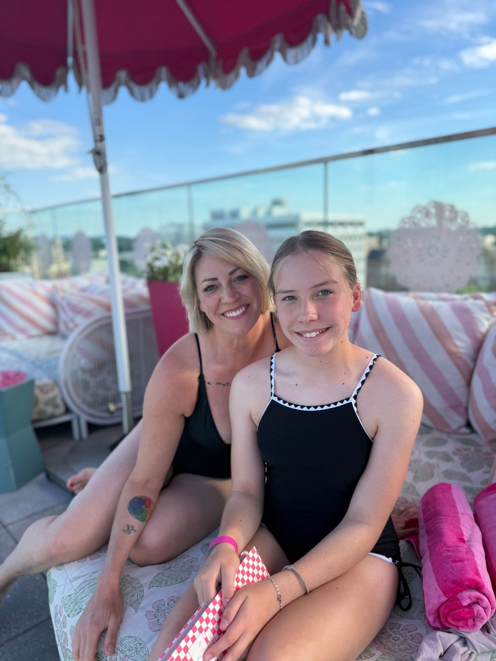 Two women in black swimsuits smile together on a rooftop pool area with pink umbrella and cushions.
