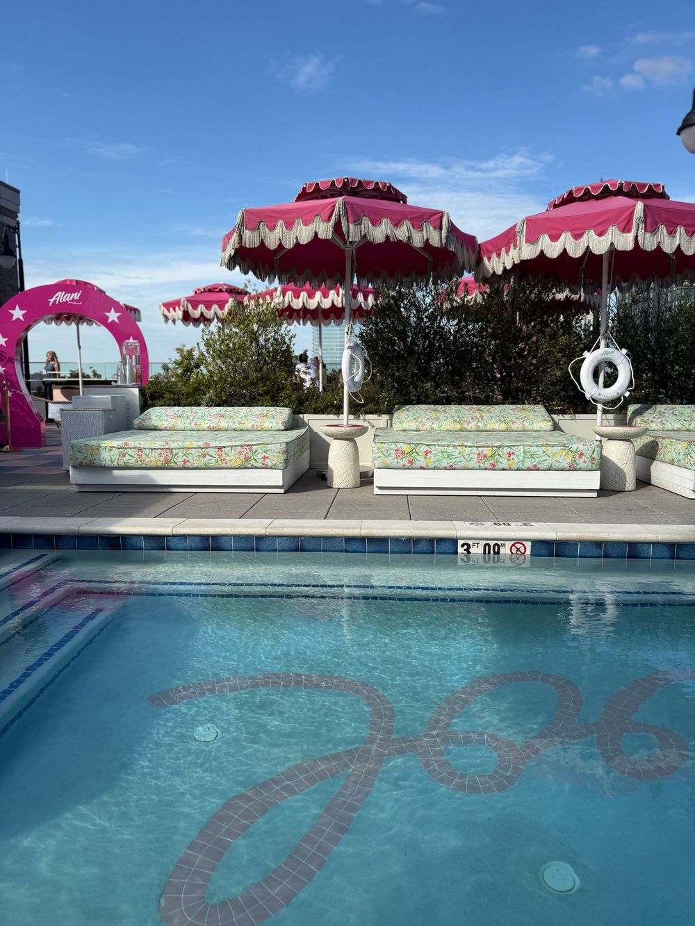 Luxury rooftop pool with pink fringed umbrellas, floral loungers, and decorative tile mosaic on a sunny day.