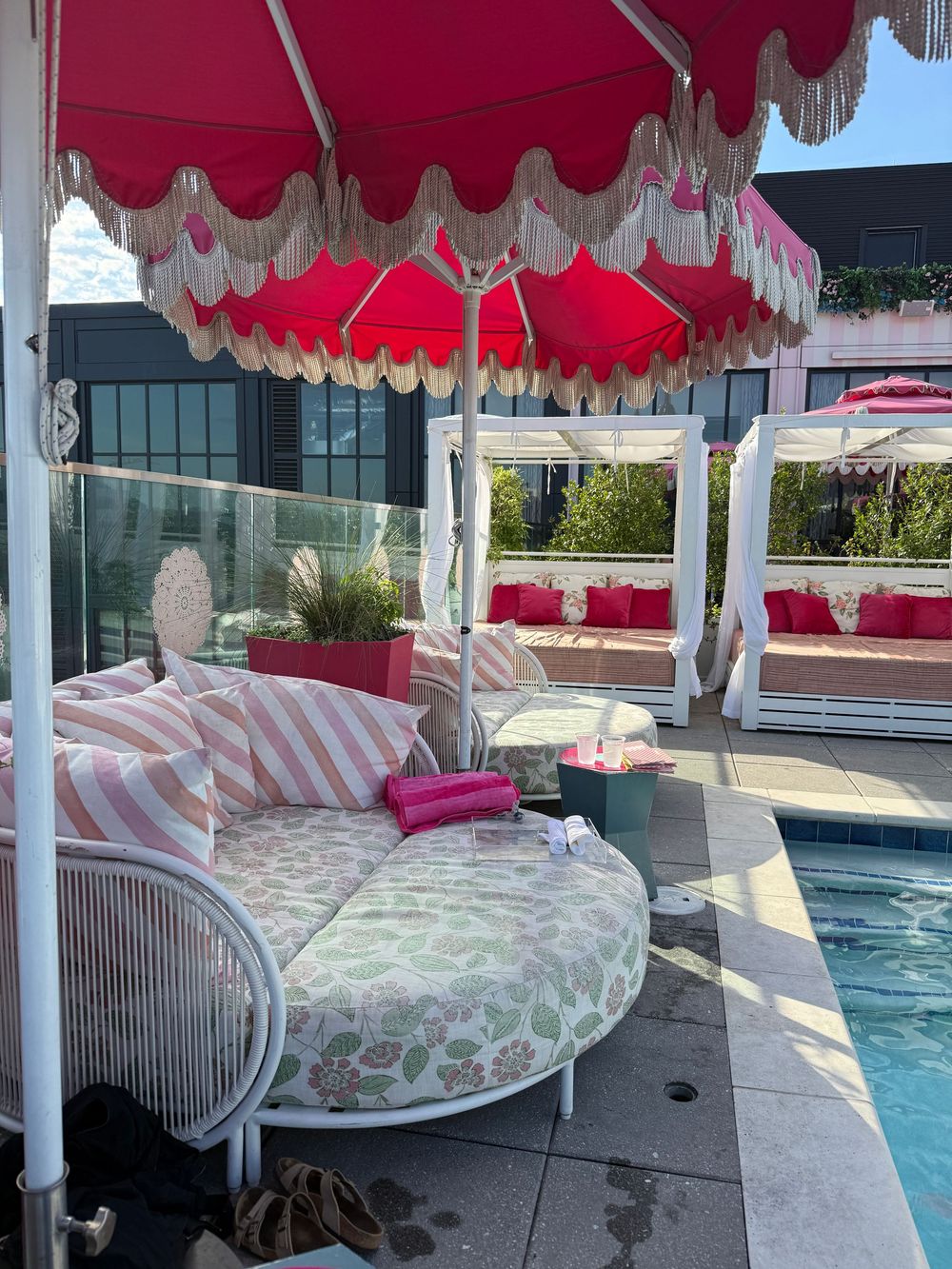Luxurious poolside cabana with round floral daybed, pink striped pillows, and vibrant red fringed umbrella at upscale resort.