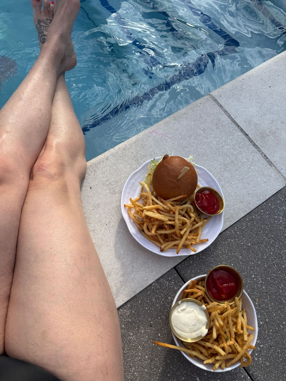 Person relaxing poolside with two plates of french fries, a burger, and ketchup on a sunny day.