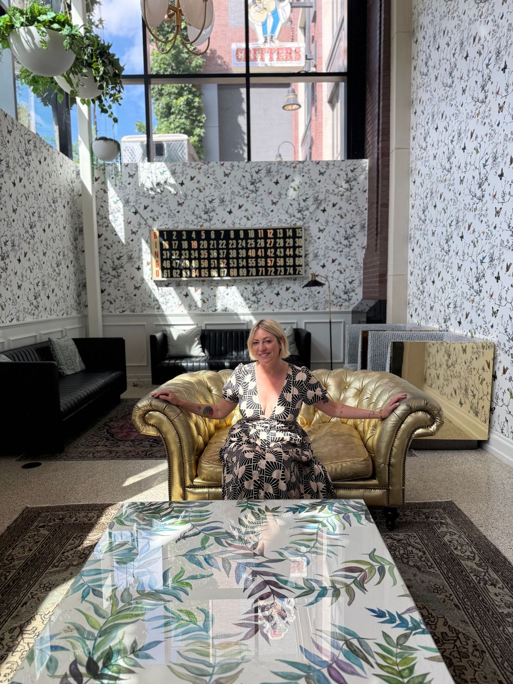 Woman in floral dress relaxing on gold velvet sofa in stylish terrazzo-walled hotel lobby with decorative coffee table.