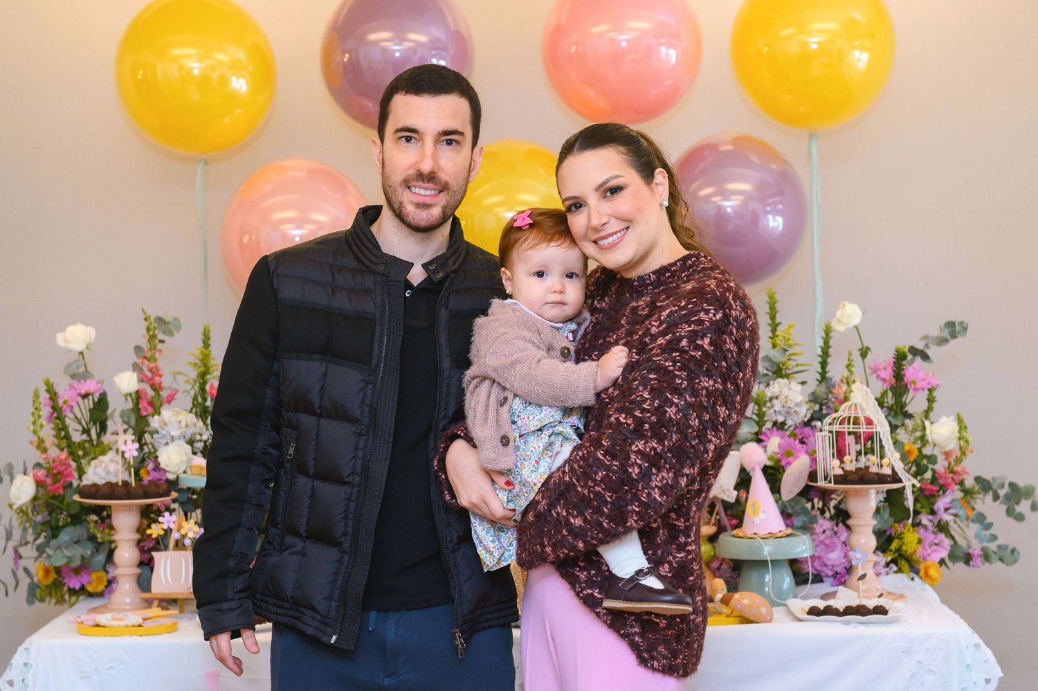 A family of three poses at a birthday party with colorful balloons and floral decorations in the background.