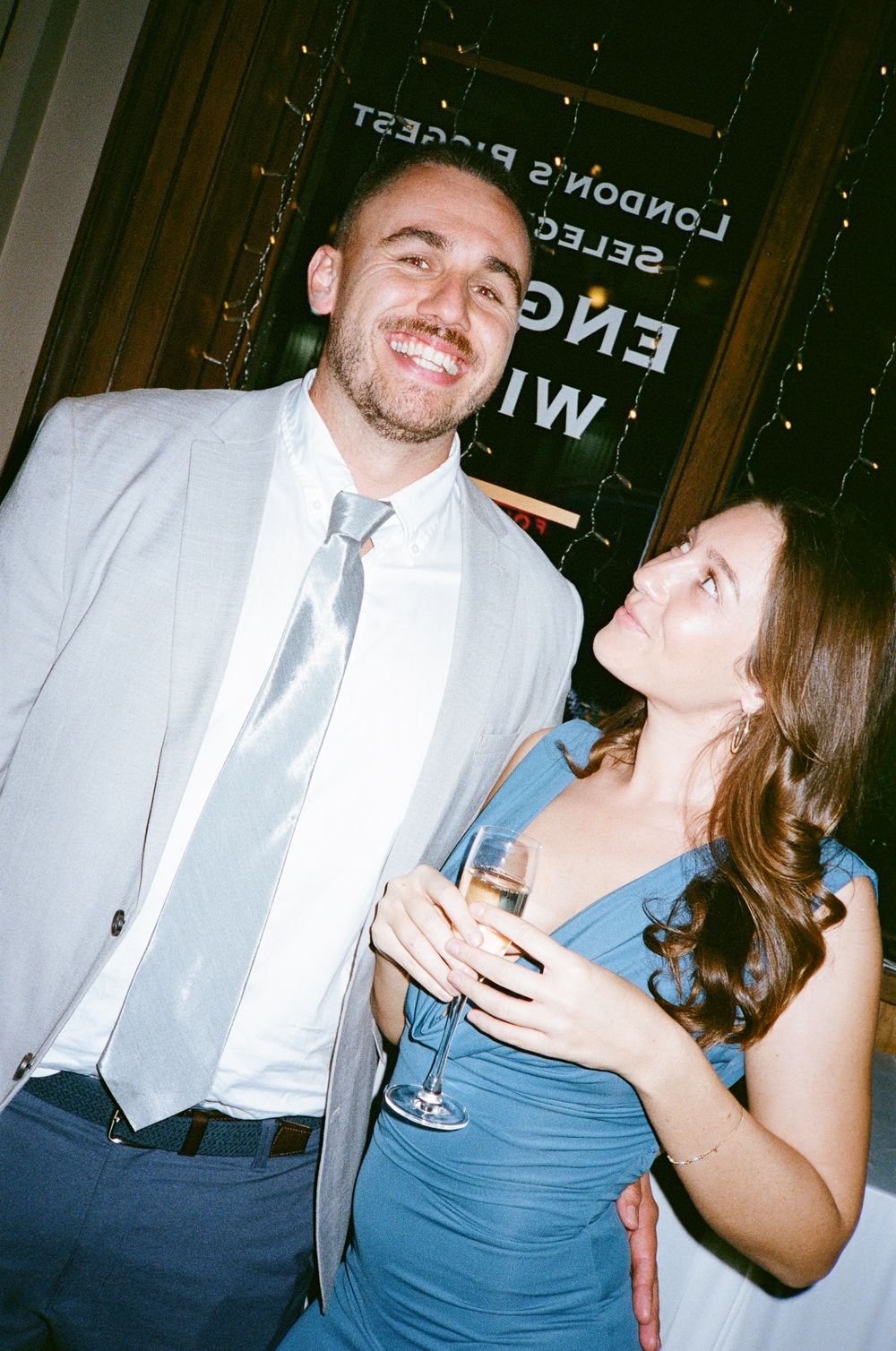 A smiling man in a grey suit and tie stands with a woman in a blue dress holding a wine glass at a formal event.