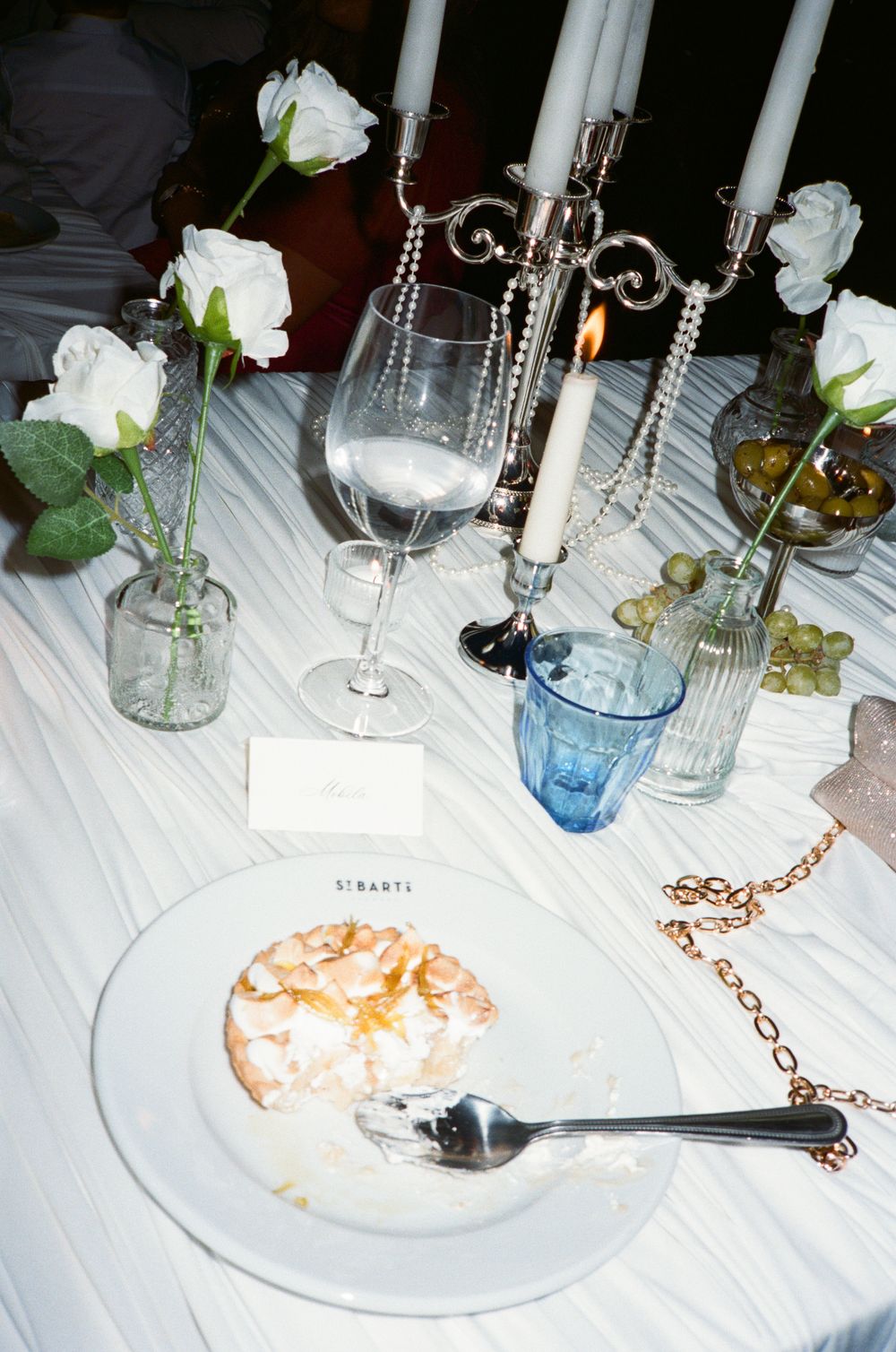 Elegant dinner table setting with white roses, candelabra, wine glass, blue tumbler, grapes, and a dessert plate with spoon.