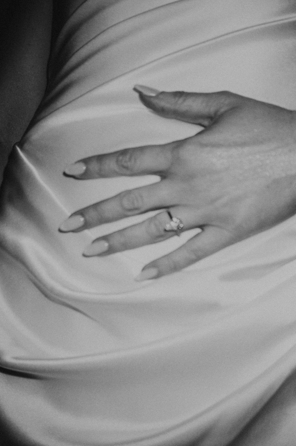 Black and white close-up of a womans hand with long nails wearing a diamond engagement ring on a satin dress.