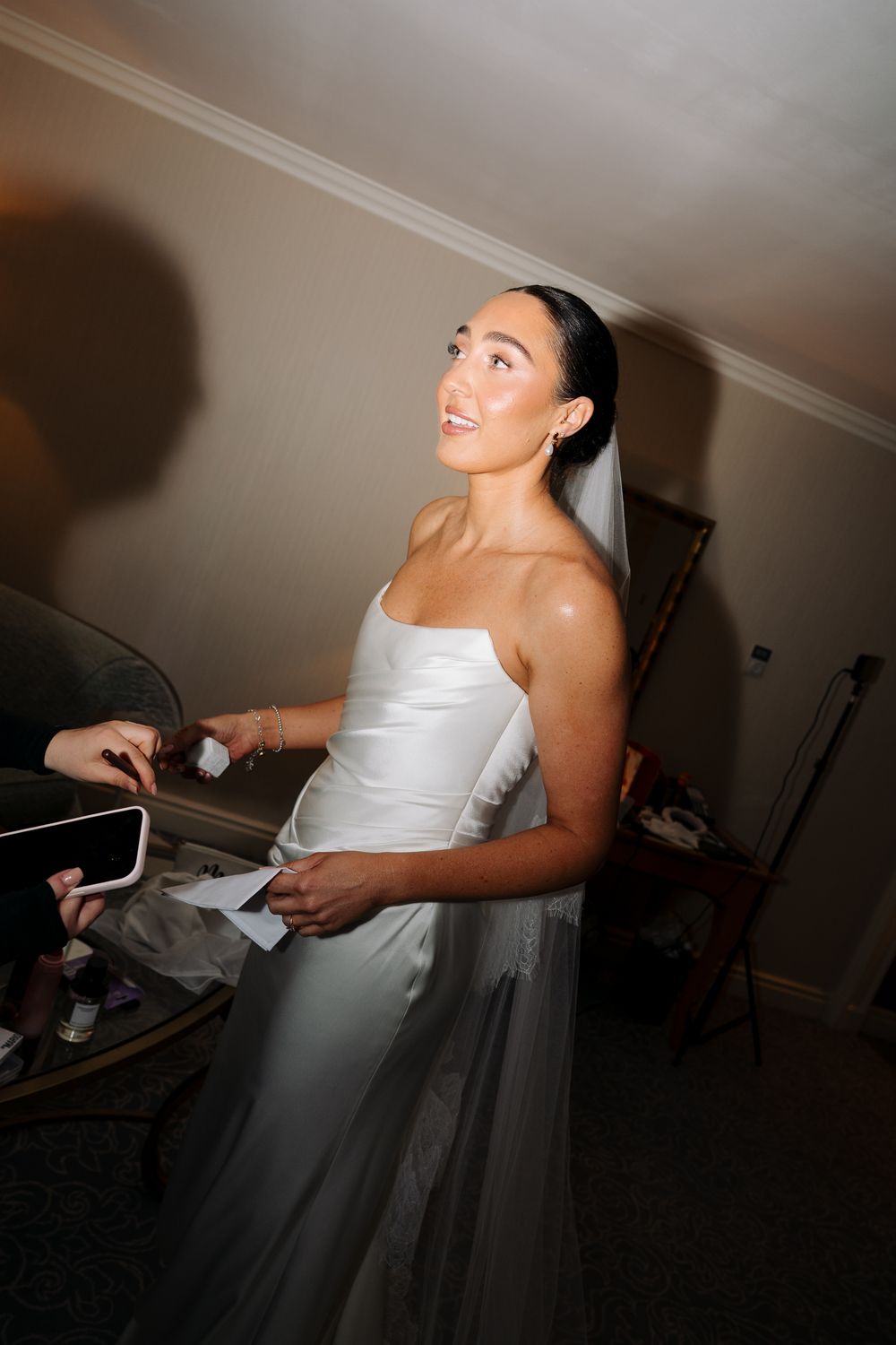A bride in a white strapless gown and veil smiles while being helped getting ready indoors.