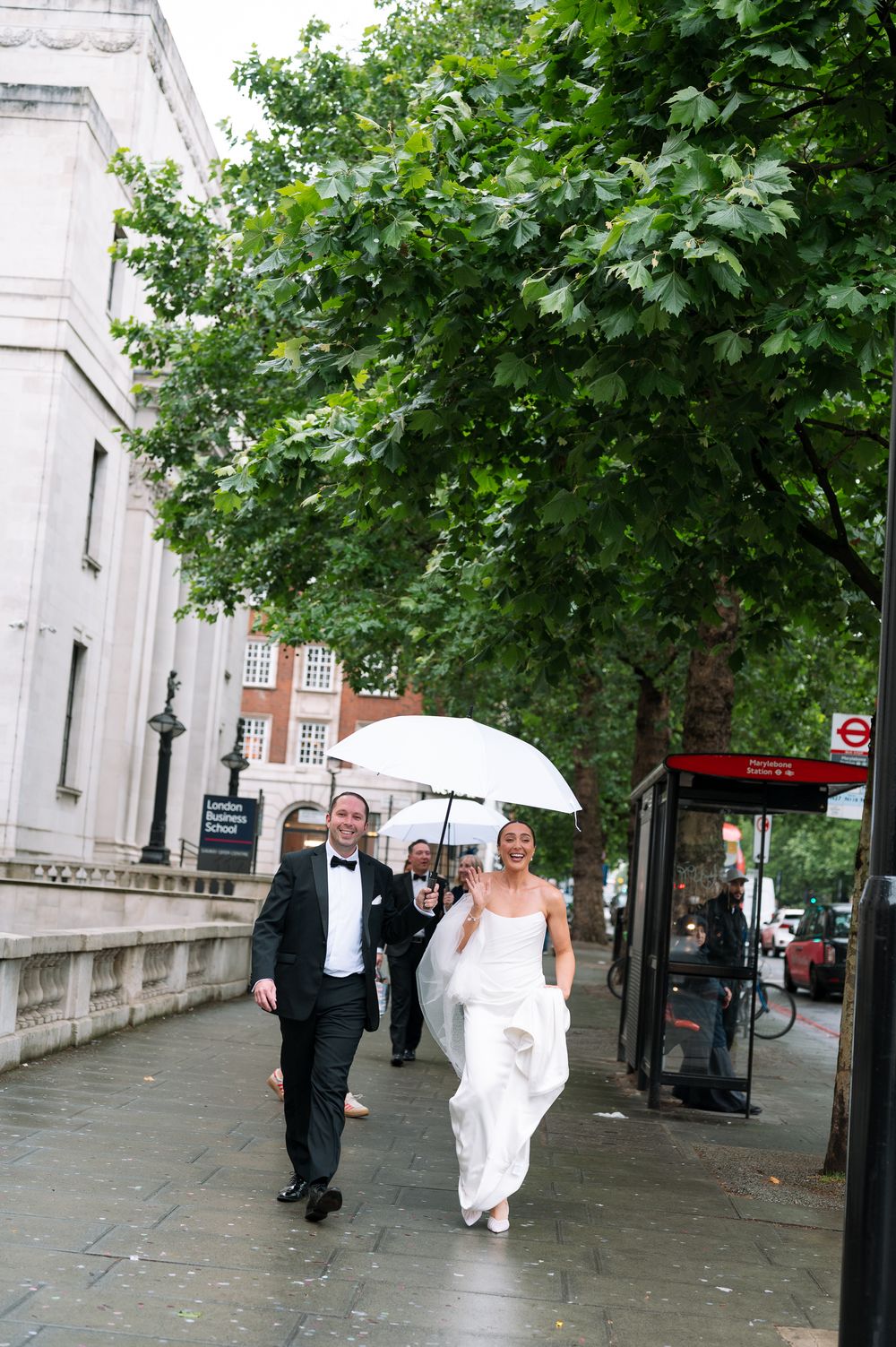 Bride and groom walking along a London street in the rain, bride holding a white umbrella, wearing wedding dress and tuxedo.