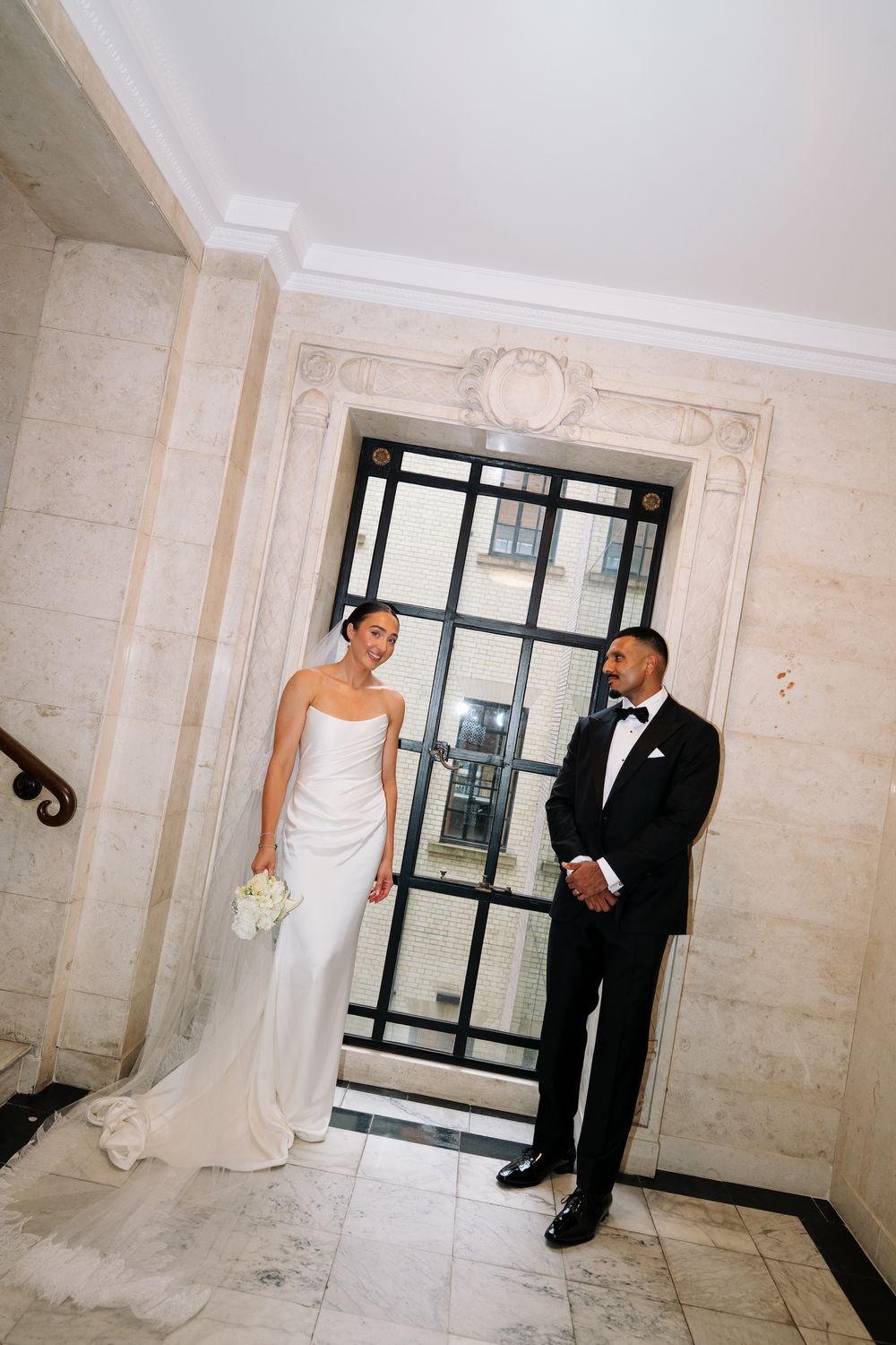 Bride in white strapless gown and groom in black tuxedo posing by ornate window in elegant marble venue.