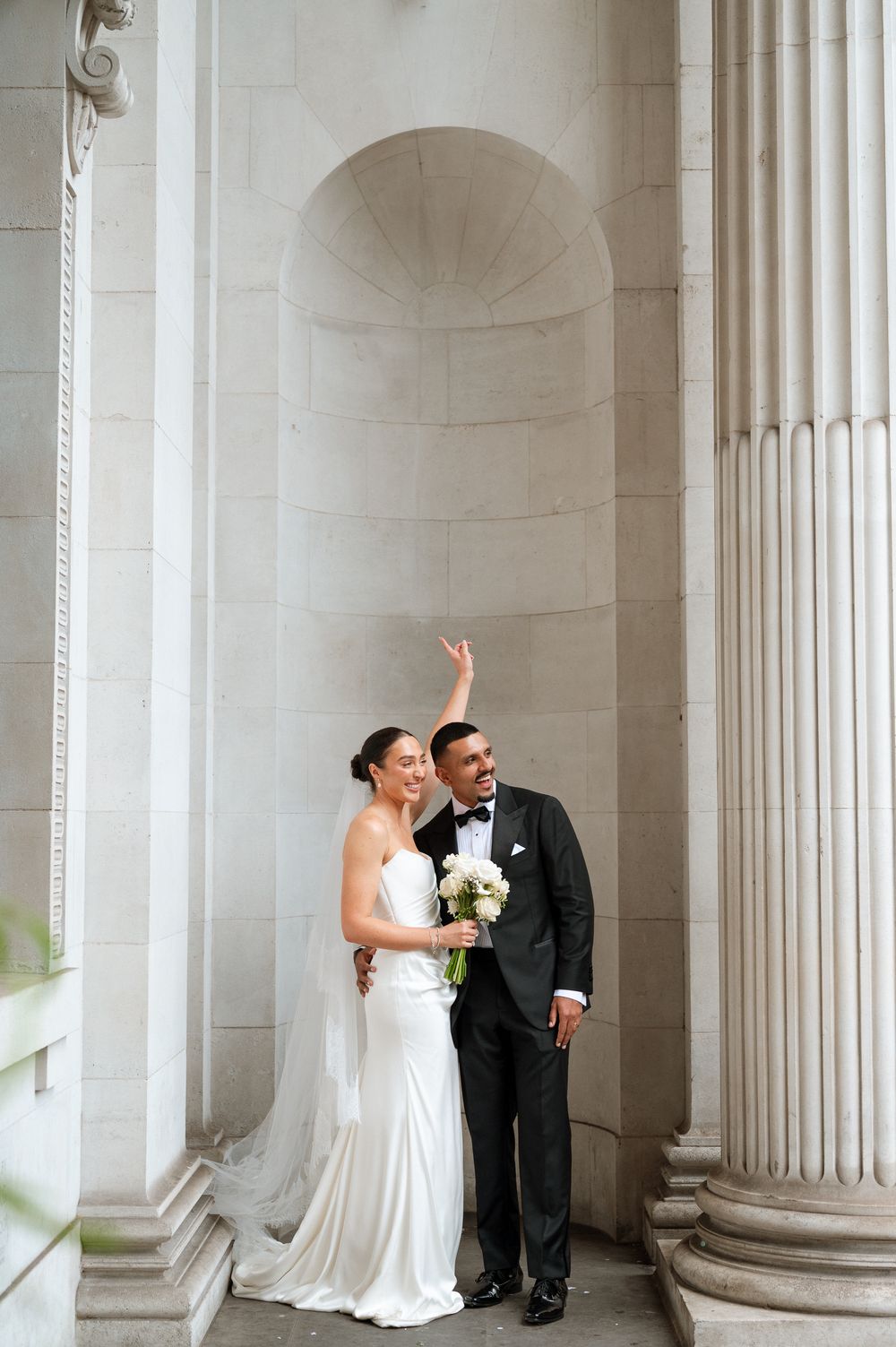 Bride in white gown and groom in black tuxedo pose by grand stone columns, groom raising hand joyfully.