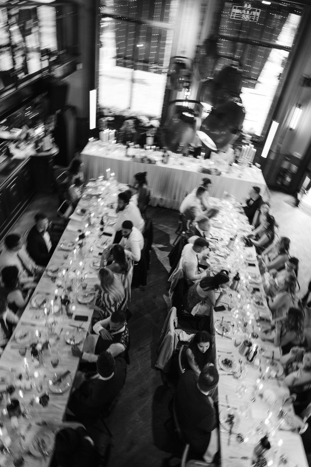Aerial black and white photo of elegant dinner party guests seated at long banquet tables in a grand restaurant.