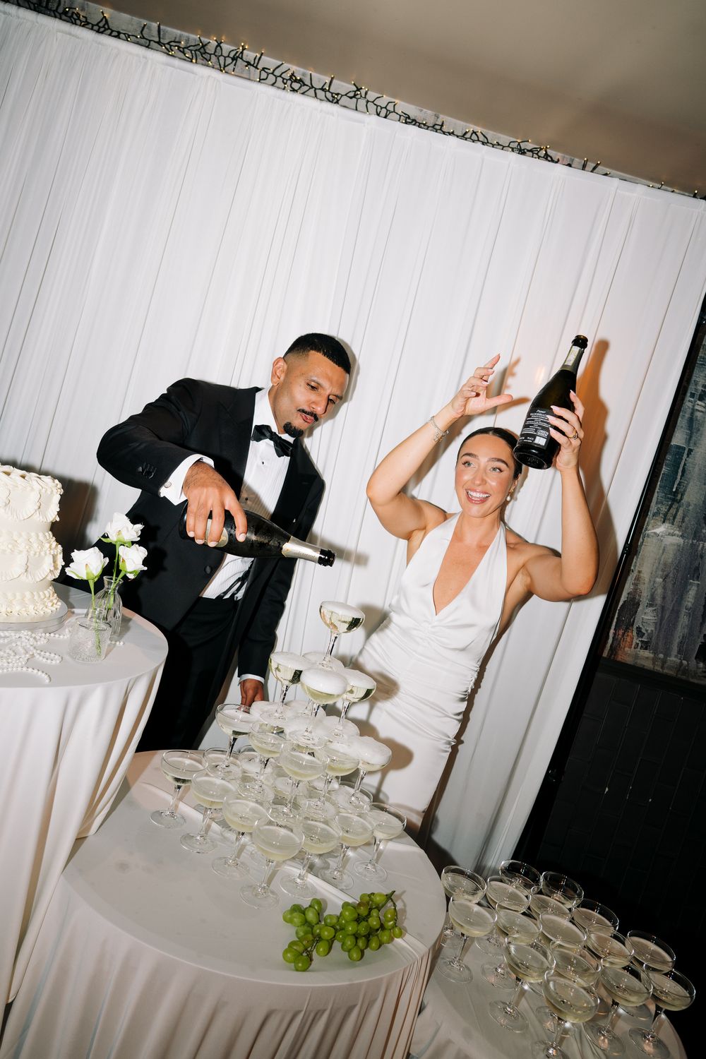 Bride and groom pouring champagne tower at wedding reception, smiling joyfully in elegant black and white attire.