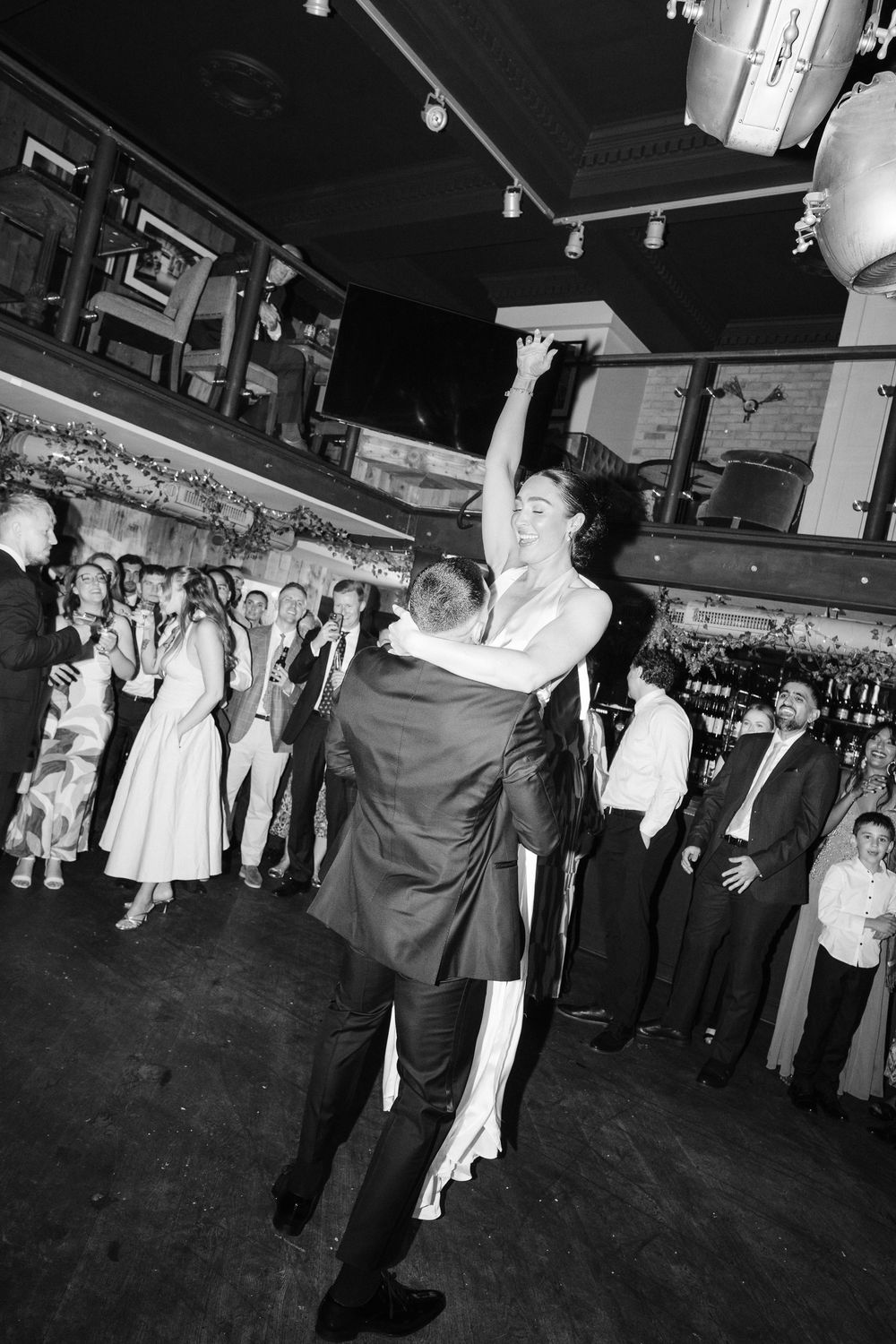 Black and white photo of a bride and groom sharing their first dance at a wedding reception with guests watching.