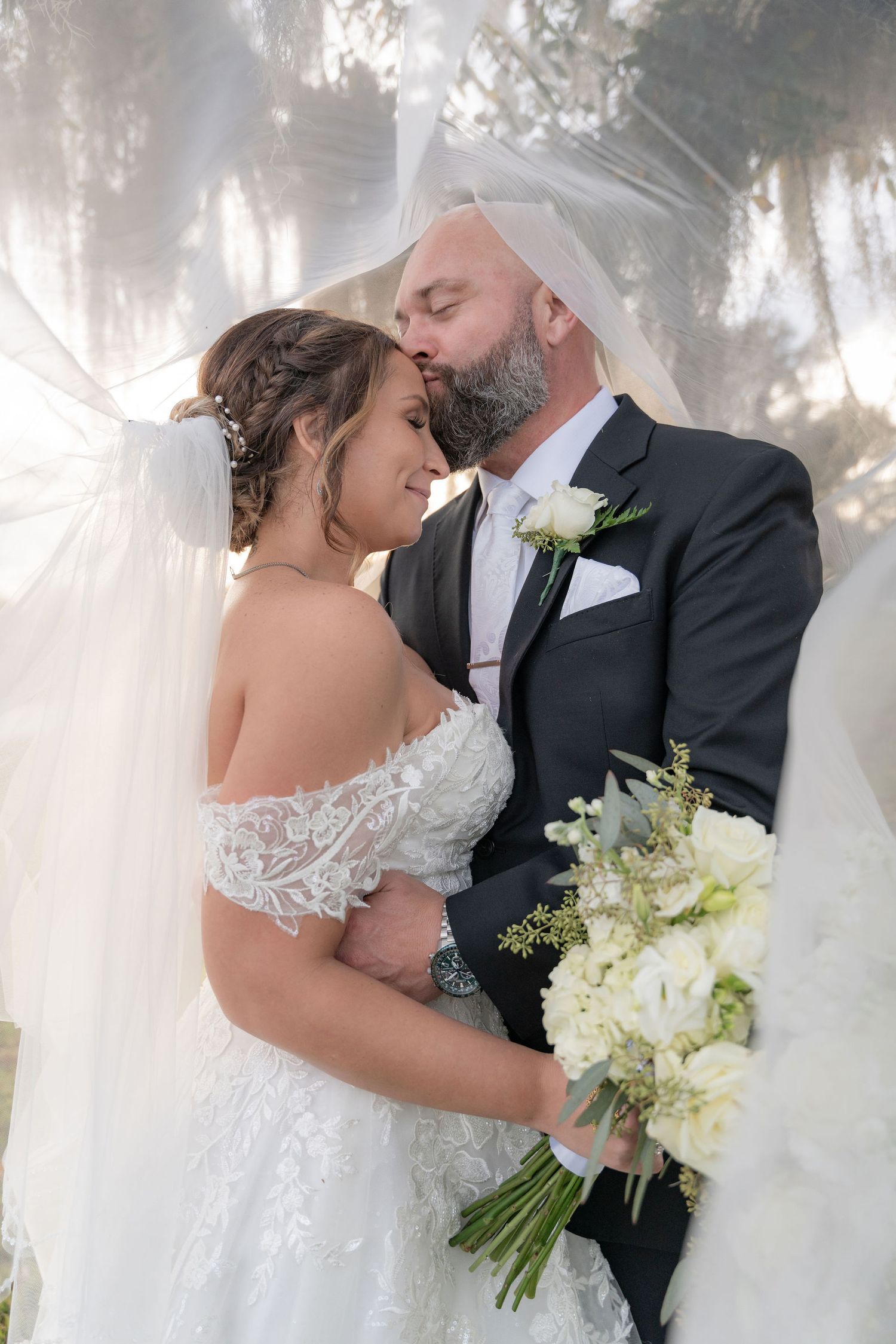 Groom kissing his bride's forehead under her veil beneath the mossy oaks at Rockin' H Ranch in Palm City, FL