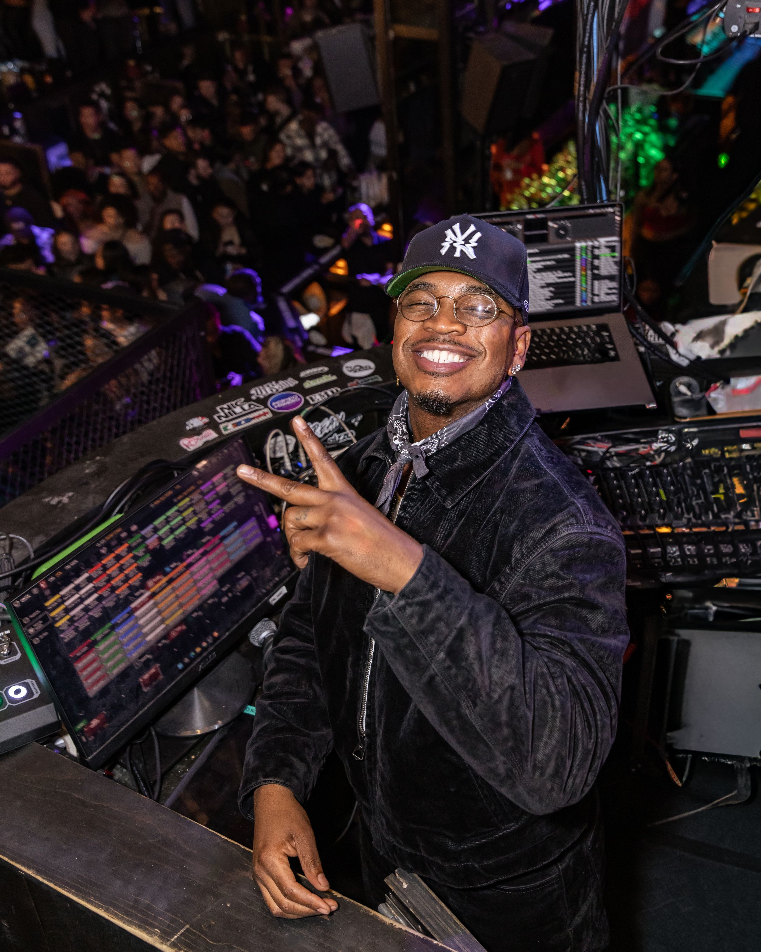 Smiling DJ in NY Yankees cap makes peace sign at DJ booth in a crowded nightclub venue.