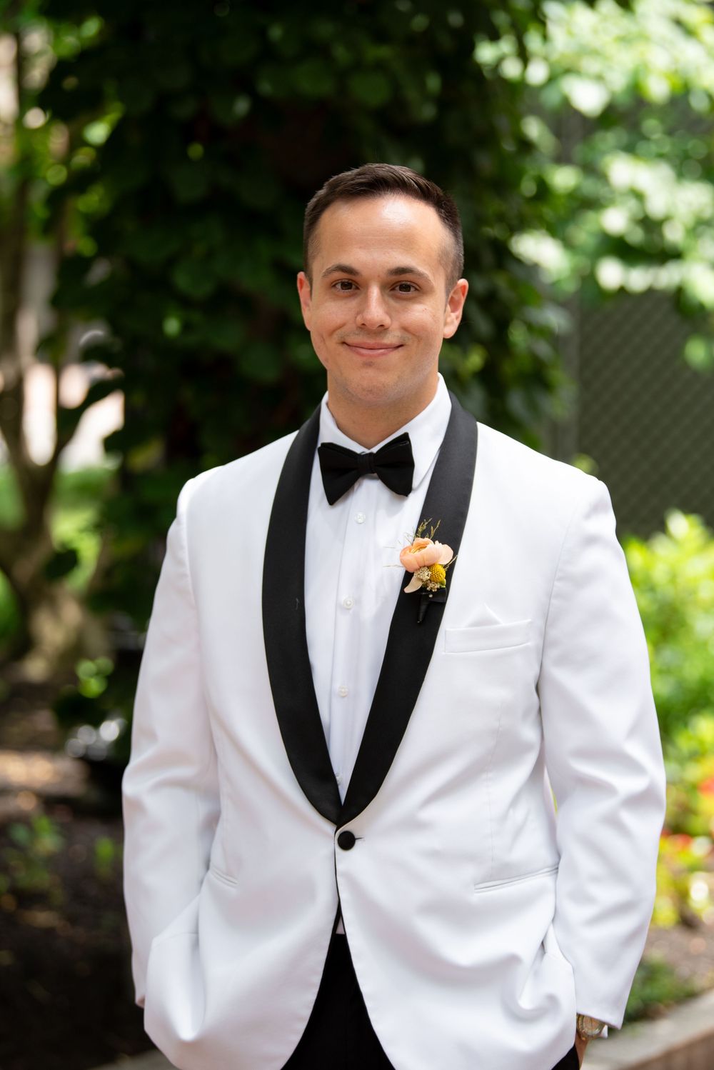 Groom in white tuxedo jacket with black lapels and bow tie wearing a floral boutonniere outdoors.