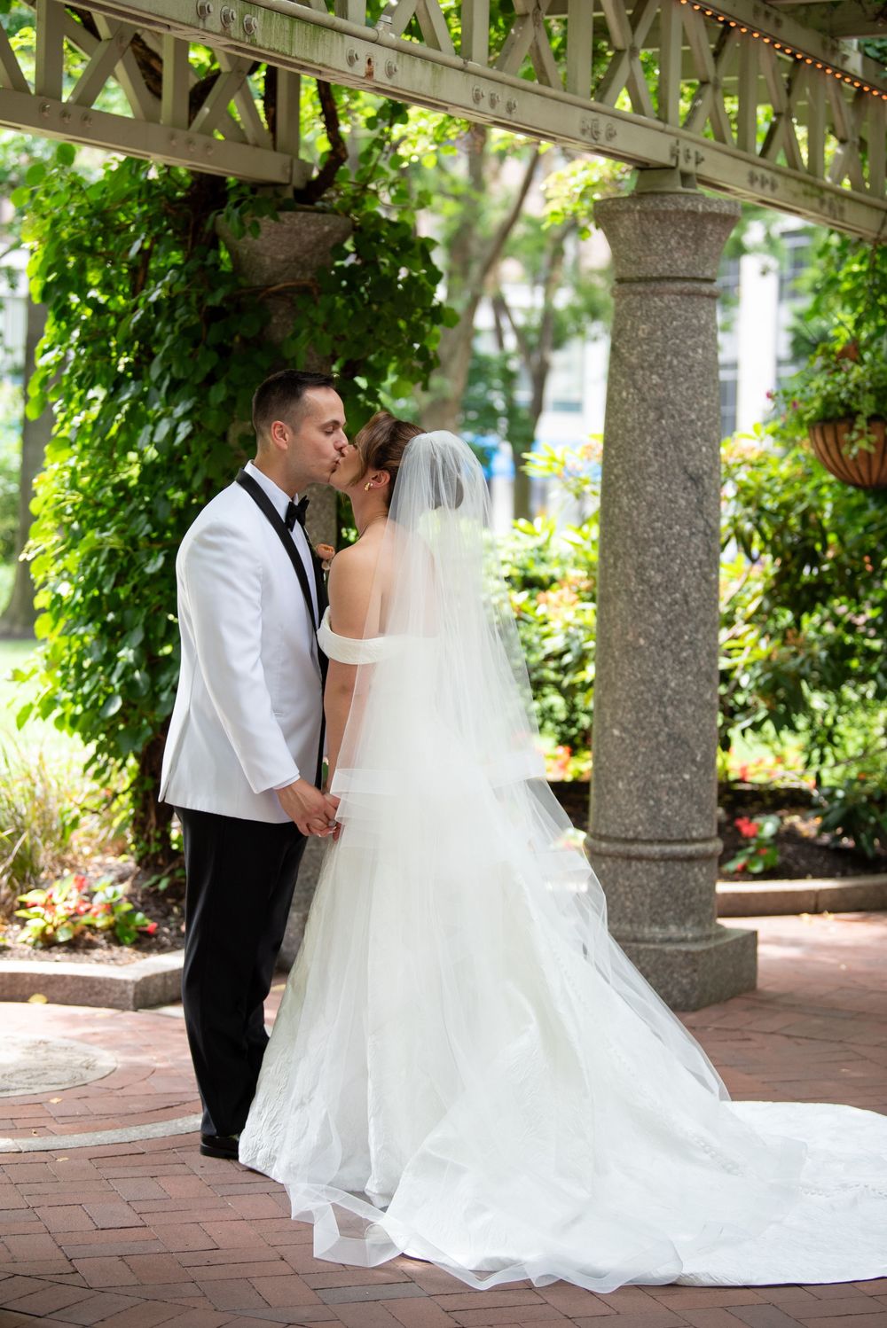 Bride and groom share a romantic kiss under a garden pergola, bride in white gown with veil and long train.