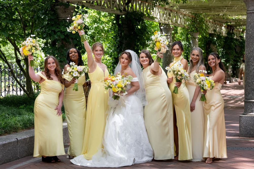 Bride in white gown poses with seven bridesmaids in yellow dresses holding bouquets outdoors under a floral arbor.