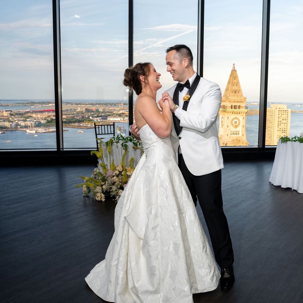 Bride and groom share first dance at elegant high-rise wedding venue overlooking Boston harbor at golden hour.
