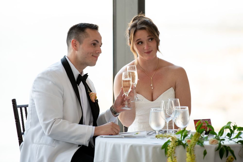 Bride and groom toasting with champagne glasses at wedding reception table, wearing formal white tuxedo and strapless gown.