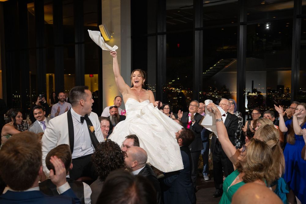 Bride laughing joyfully while being lifted by guests during lively wedding reception celebration indoors at night.