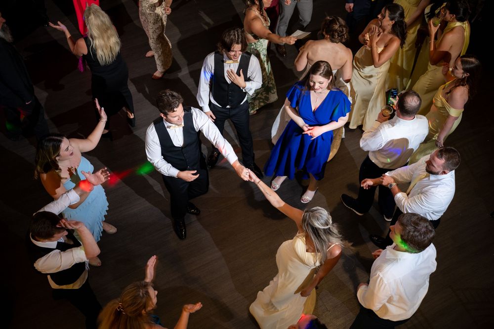 Guests dancing at a lively wedding reception, with a couple in blue dress and suit at center.