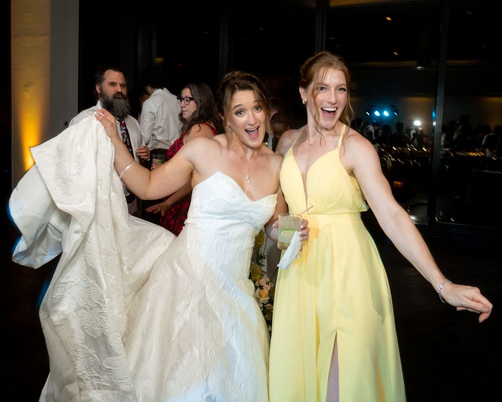 Bride in white gown and bridesmaid in yellow dress laughing joyfully at a wedding reception.