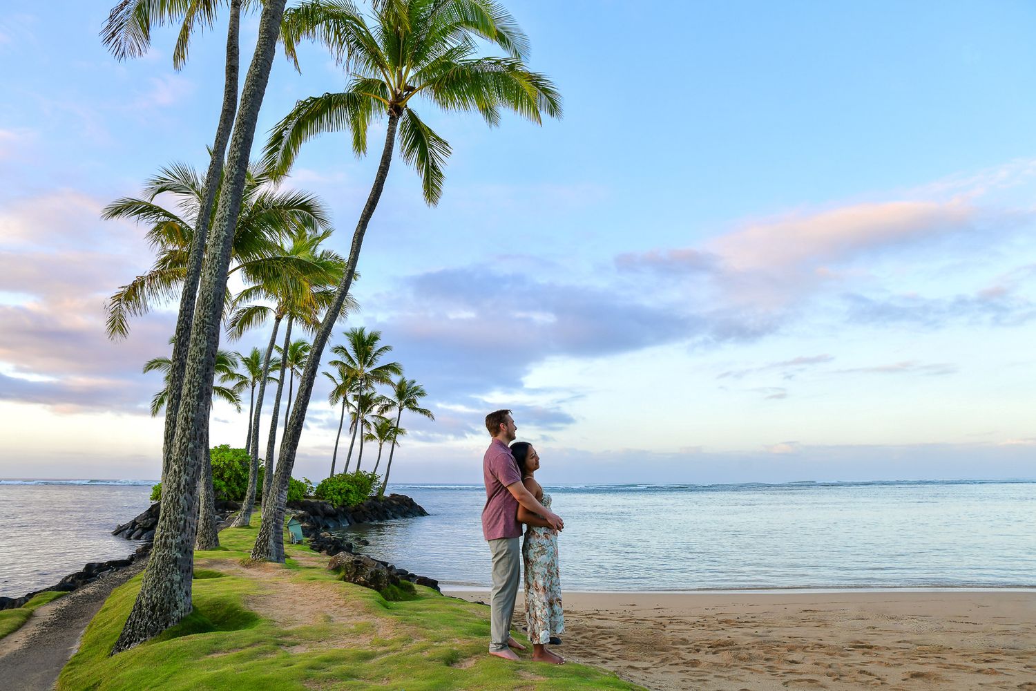 Couple embracing on a tropical beach at sunset with tall palm trees and calm ocean waters in Hawaii.