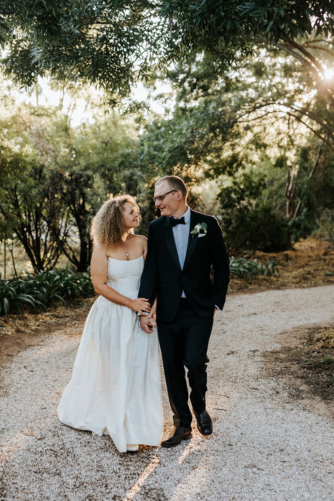 Bride and groom walking hand in hand on a garden path, sharing a loving glance in golden hour light.