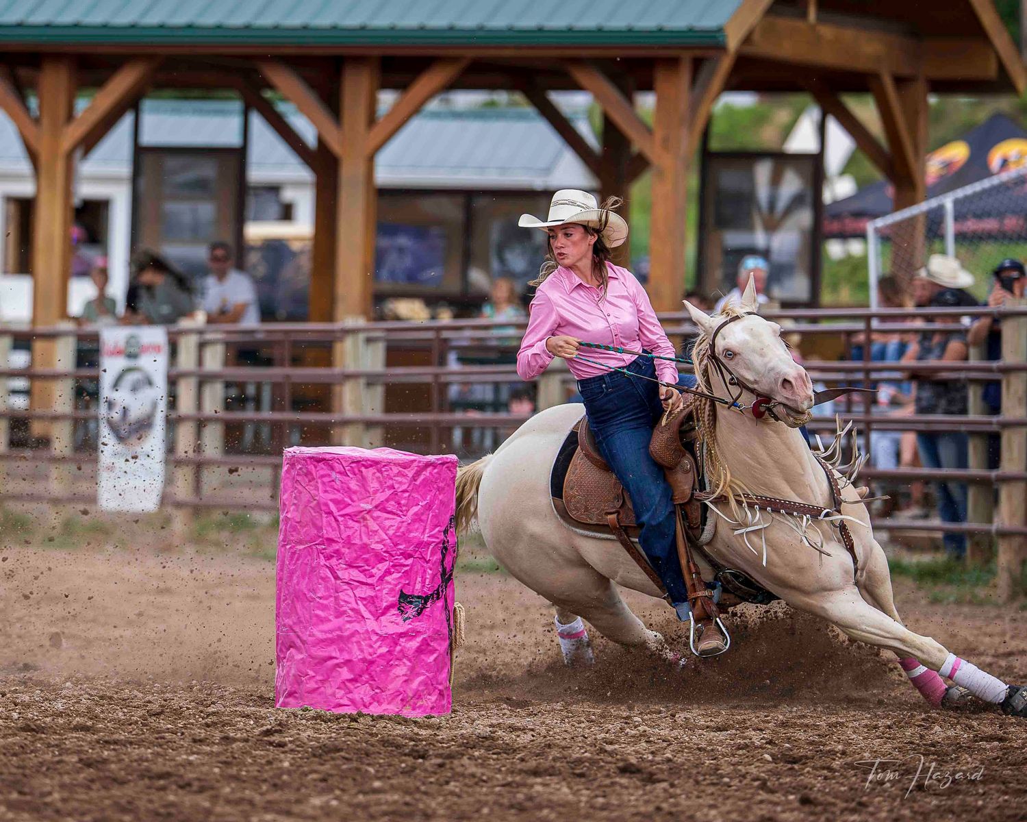 Woman in pink shirt rides white horse around pink barrel in a rodeo barrel racing competition.