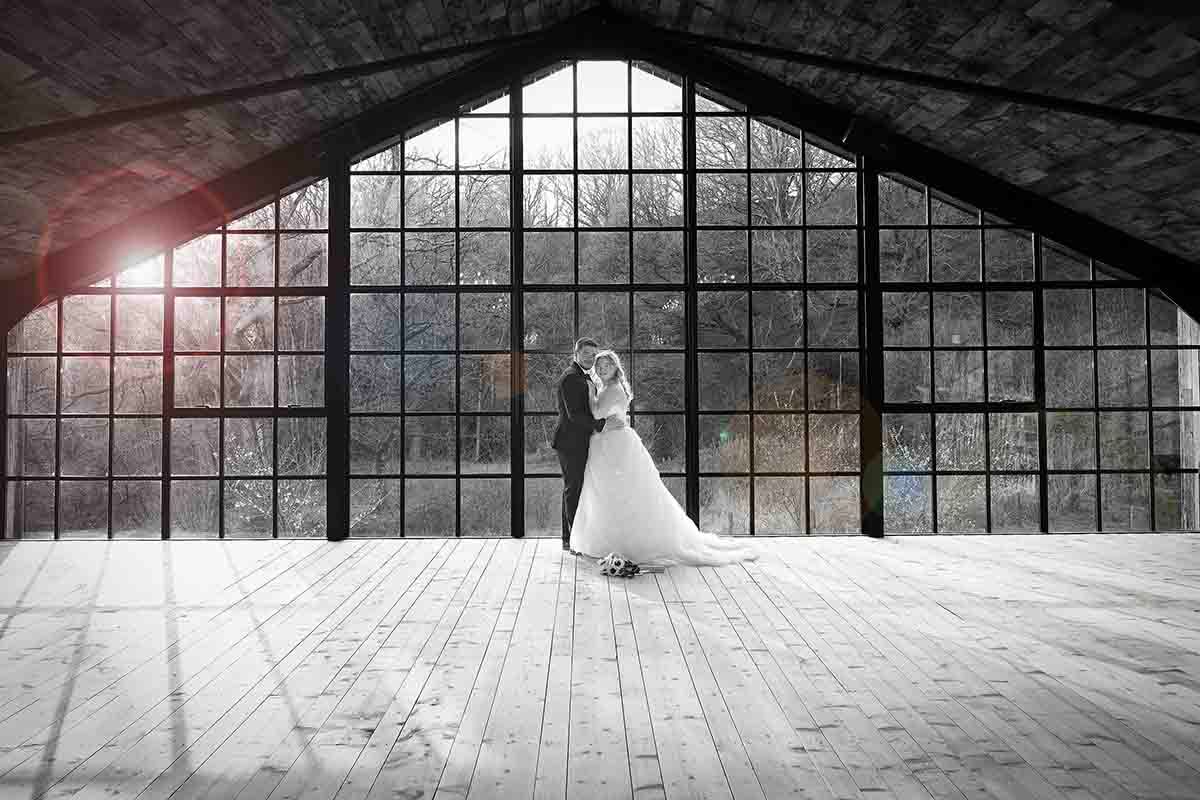 Bride and groom embracing in a grand industrial hall with large arched windows and wooden floors.