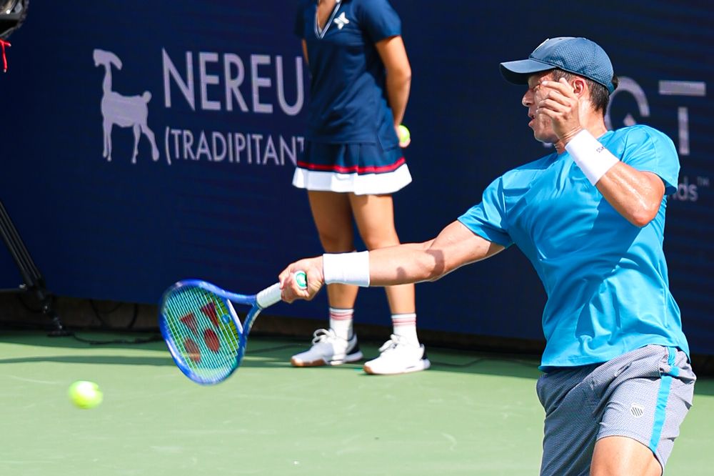 Tennis player in blue outfit hits a backhand shot on an outdoor hard court during a professional tournament match.