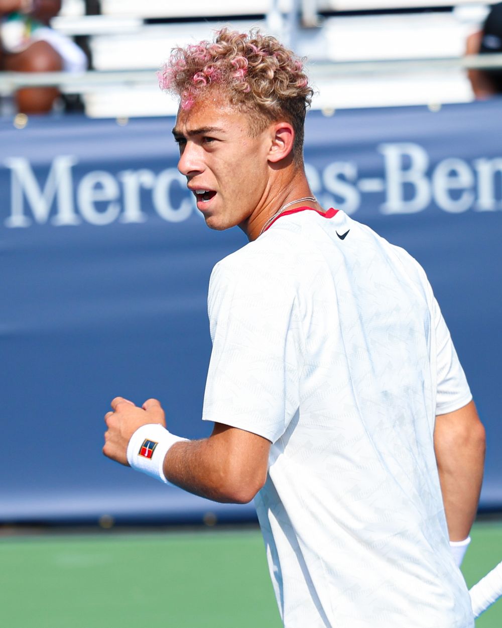Tennis player with pink curly hair wearing white Nike outfit pumps fist in celebration at Mercedes-Benz sponsored court.