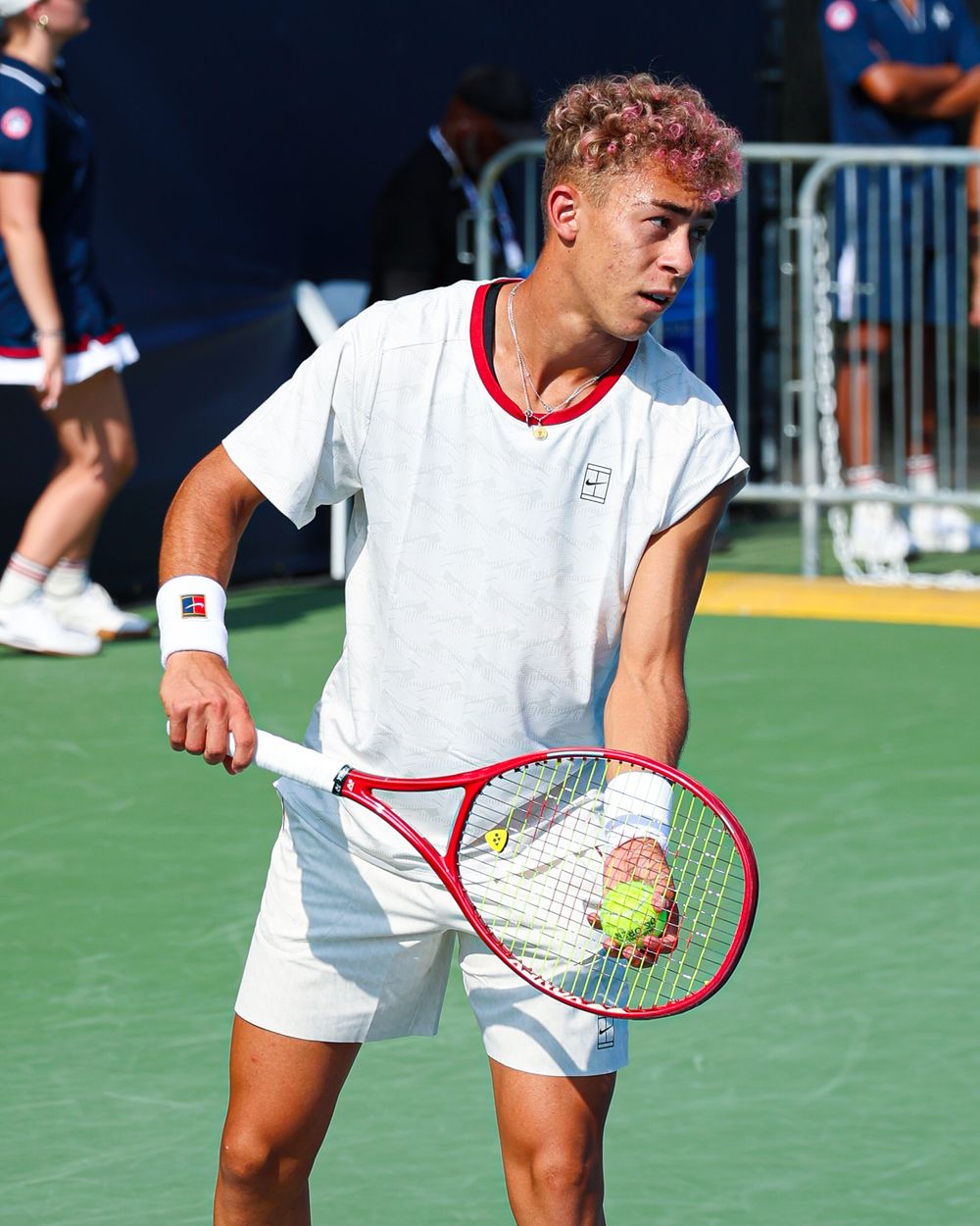 Young male tennis player with pink hair holding a racket and ball, wearing white outfit on outdoor court.