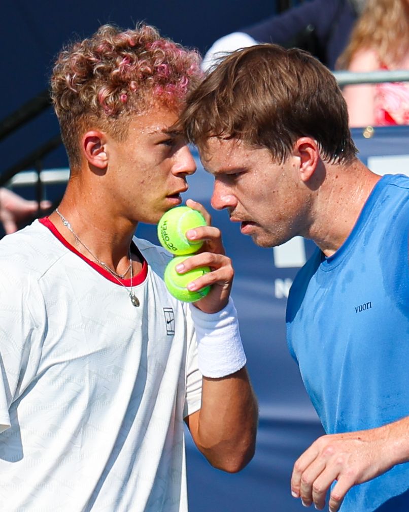 Two tennis players in sportswear huddle closely, one holding a yellow tennis ball, discussing strategy on a sunny court.