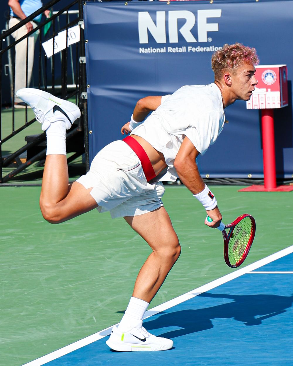Tennis player in white outfit serves on blue court at NRF National Retail Federation sponsored tournament.