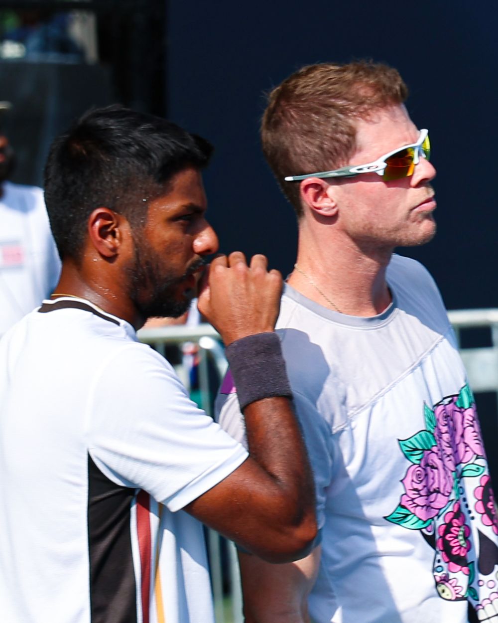 Two male cricket players in white jerseys conversing outdoors, one wearing white sunglasses at a sports event.