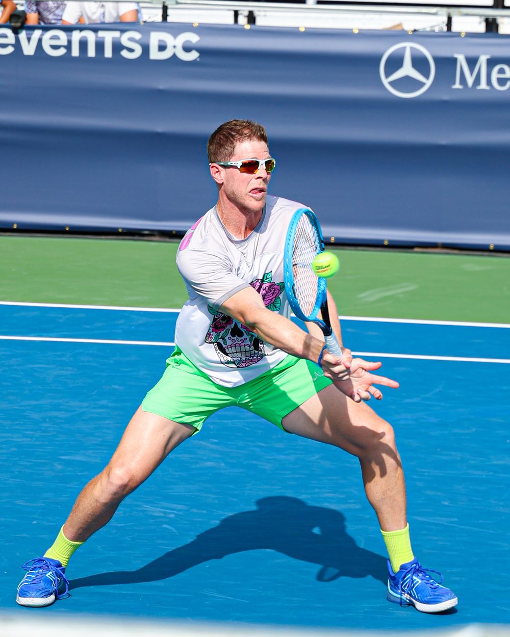 Man in neon green shorts and sunglasses playing tennis on a blue court at an Events DC tournament.