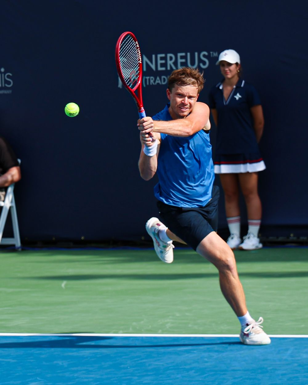 Male tennis player in blue shirt executing a backhand shot on outdoor court during competitive match.